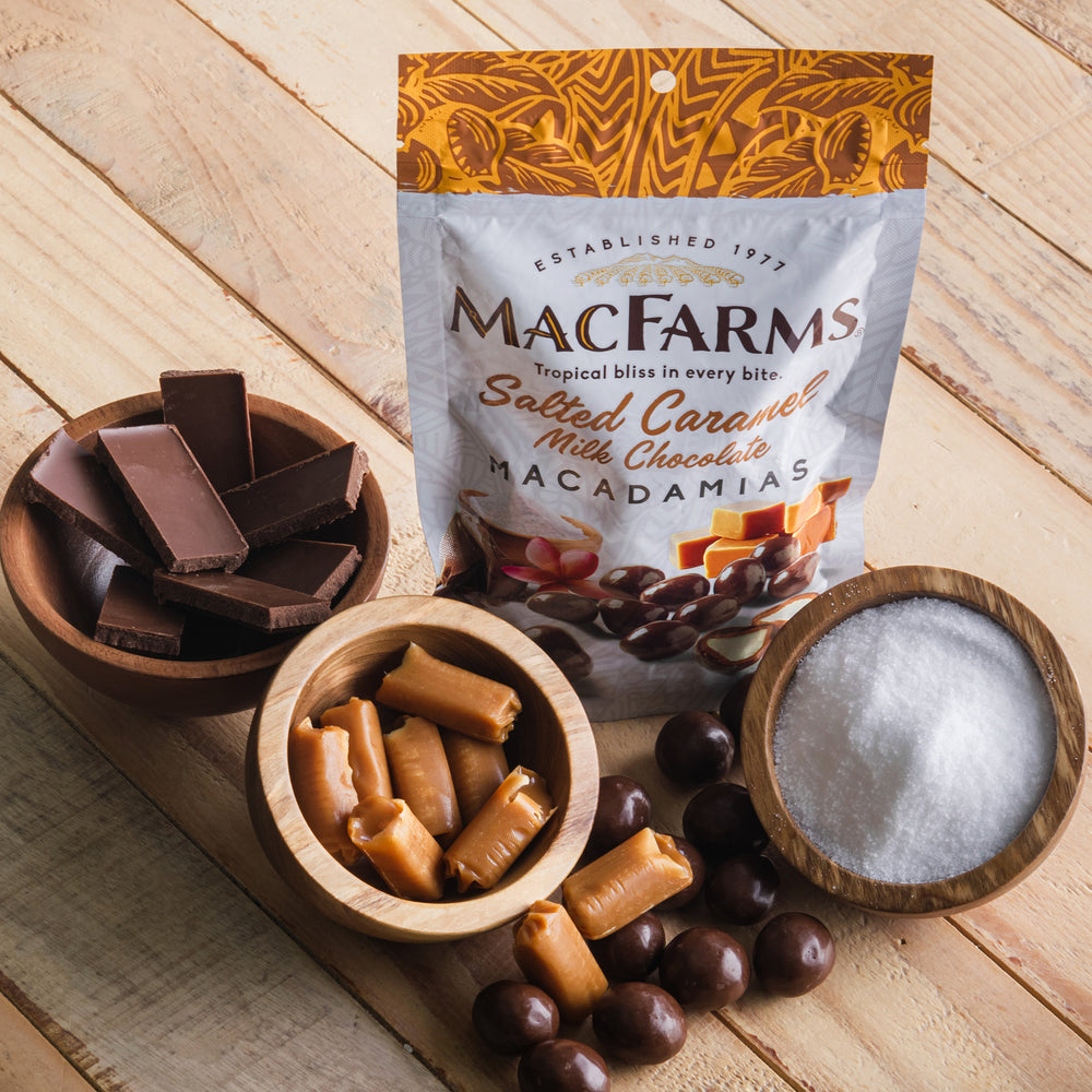 
            
                Load image into Gallery viewer, salted caramel milk chocolate macadamias - MacFarms on wood table by bowls of chocolate, caramel, and salt
            
        