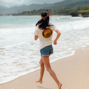 back of woman running toward ocean wearing vintage white t-shirt with burlap bag and macadamias design 
