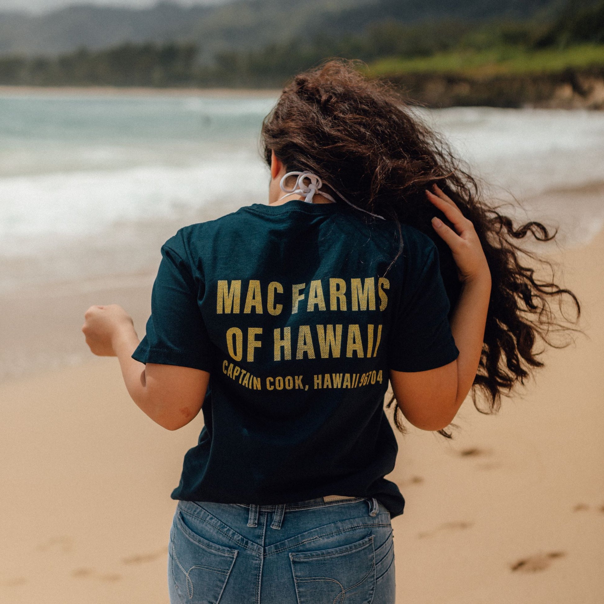back of woman walking on beach with navy tee "mac farms of hawaii" on it