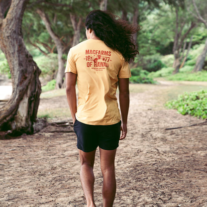 back of man walking in trees with mustard t-shirt on with "macfarms of hawaii 1977" and truck graphic 